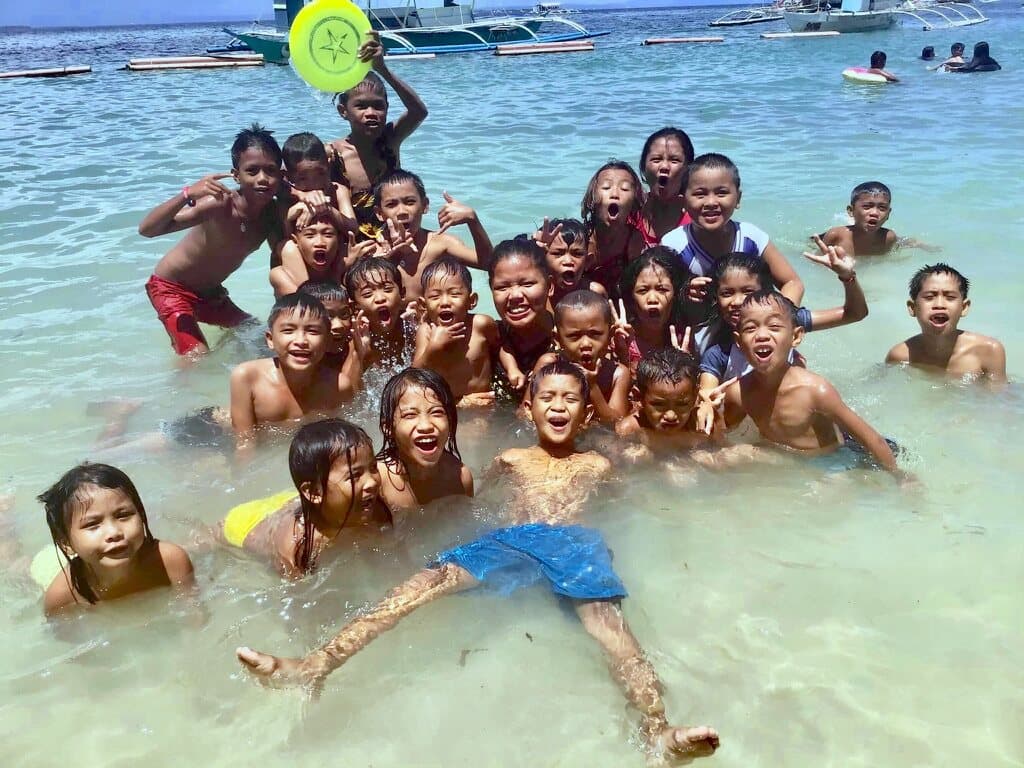 A crowd of children in the sea on a beach day, cheering at the camera.