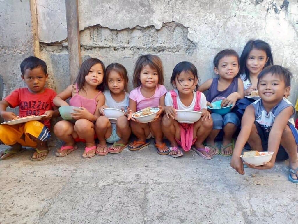 Small children sitting in a row against a wall, each with a bowl of food.