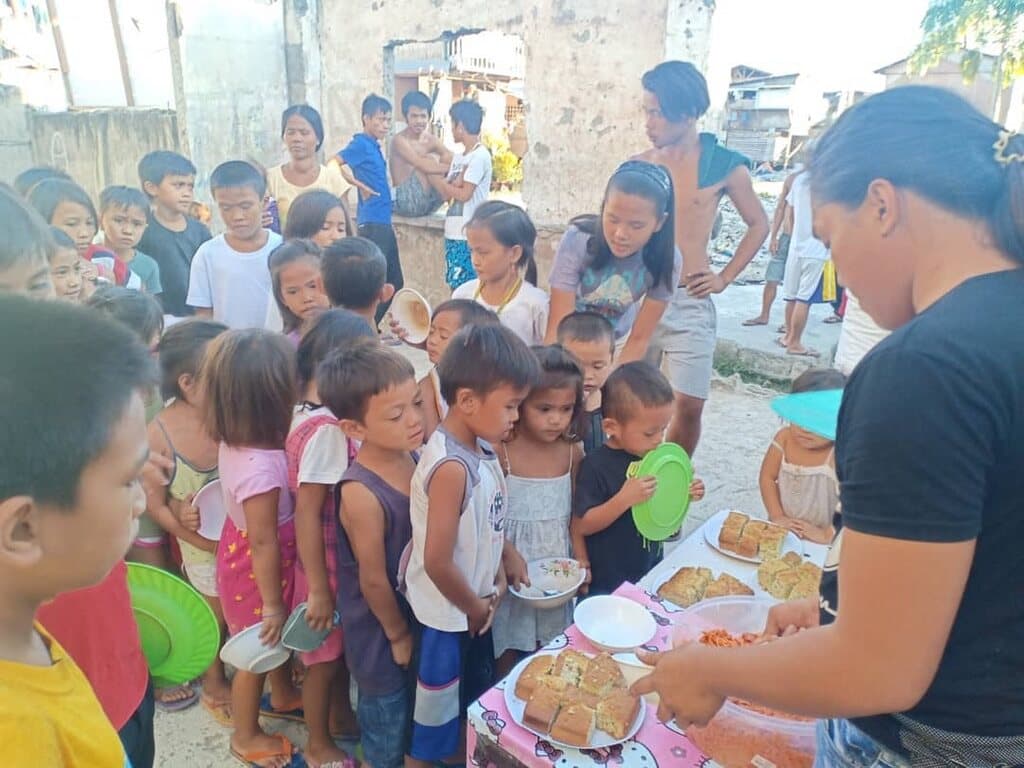 Children crowding in a line with plates while a volunteer serves food.