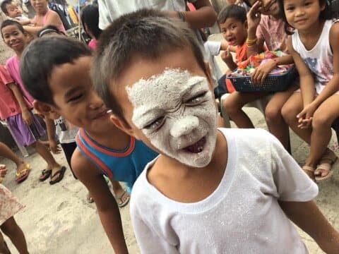 A boy with his face covered in white powder from a game, laughing, with other kids behind him.