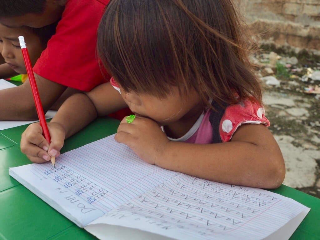 A little girl bent over her notebook, practising letters with a red pencil.
